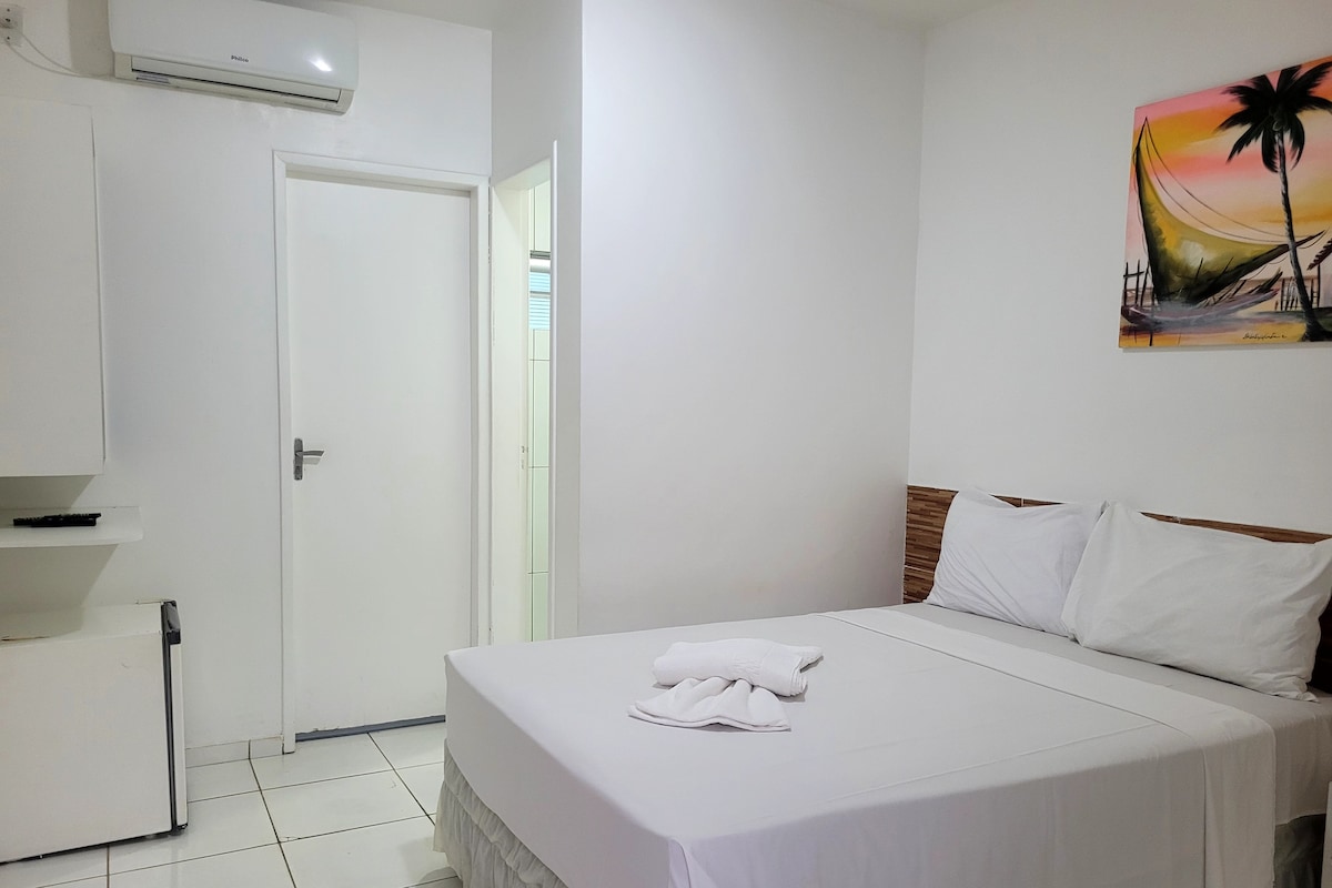 A hotel room with a bed, a white wall, a painting, and a door.