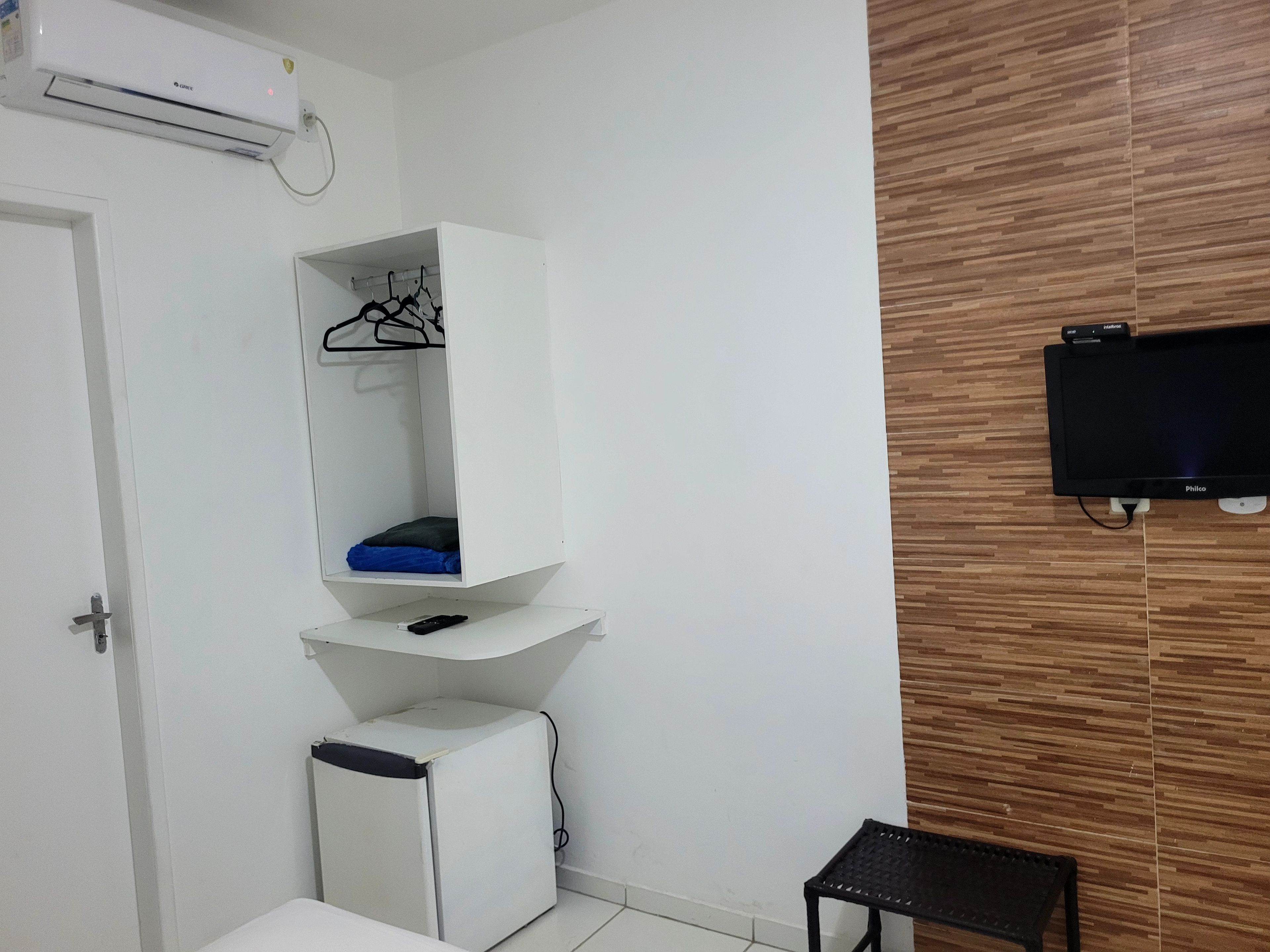 A room with a white wardrobe, a small white refrigerator, a black TV mounted on the wall, and a wooden panel design on the wall.