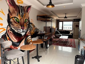 Una sala de estar con un gran mural de un gato, un sofá, una mesa de comedor y una zona de cocina.