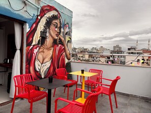 Una terraza en la azotea con sillas rojas, una mesa amarilla y un mural de una mujer.