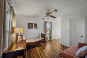 Quarto com cama, mesa de cabeceira, abajur, ventilador de teto e janela com cortinas.
