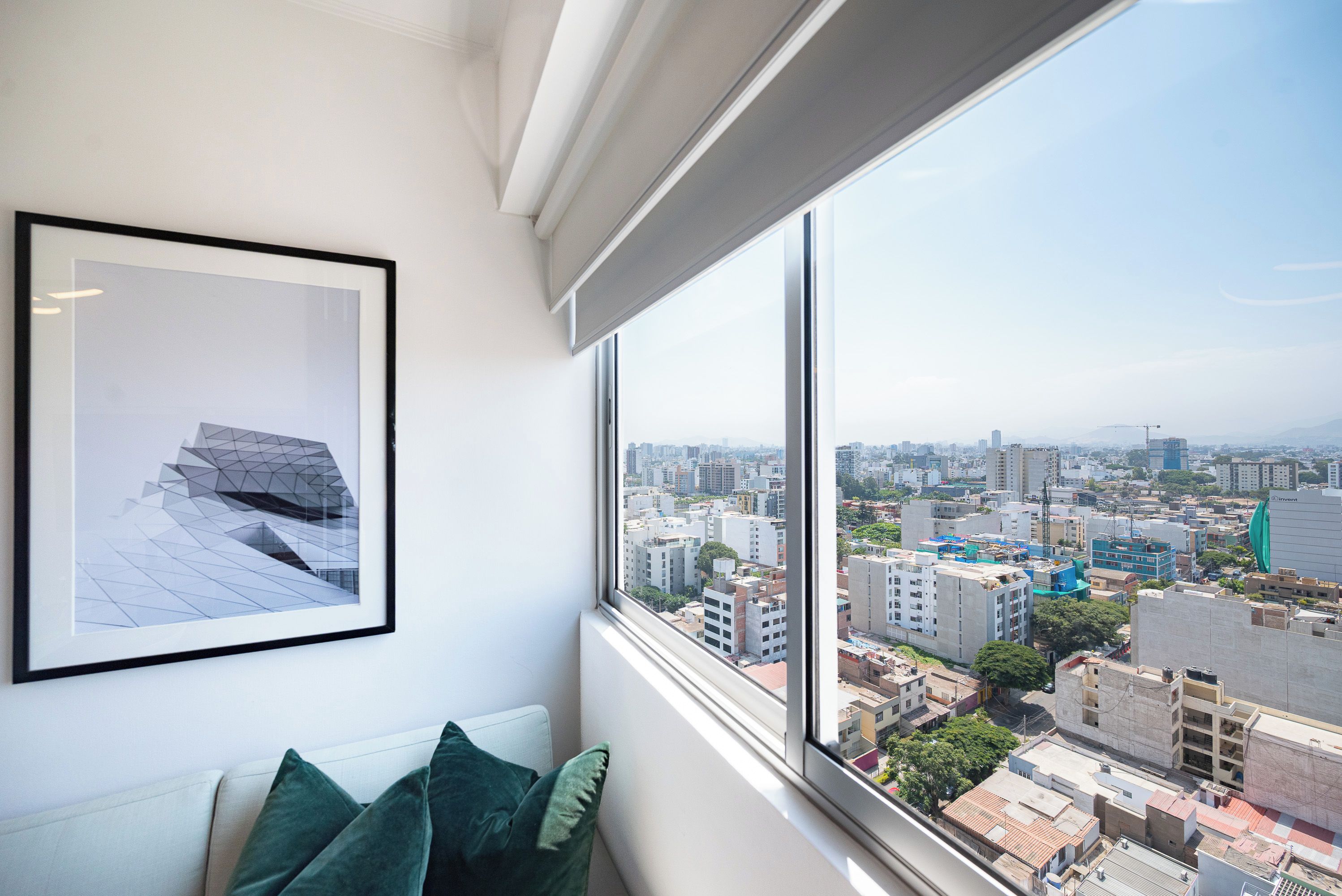 Ein Zimmer mit großem Fenster und Blick auf die Stadtlandschaft, einem gerahmten geometrischen Kunstwerk an der Wand und einem weißen Sofa mit grünen Kissen.