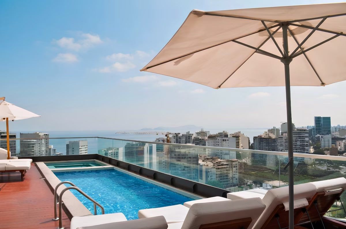 Una piscina infinita en la azotea con sillones de descanso y una sombrilla, con vistas a un paisaje urbano.