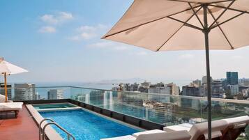 Una piscina infinita en la azotea con sillones de descanso y una sombrilla, con vistas a un paisaje urbano.