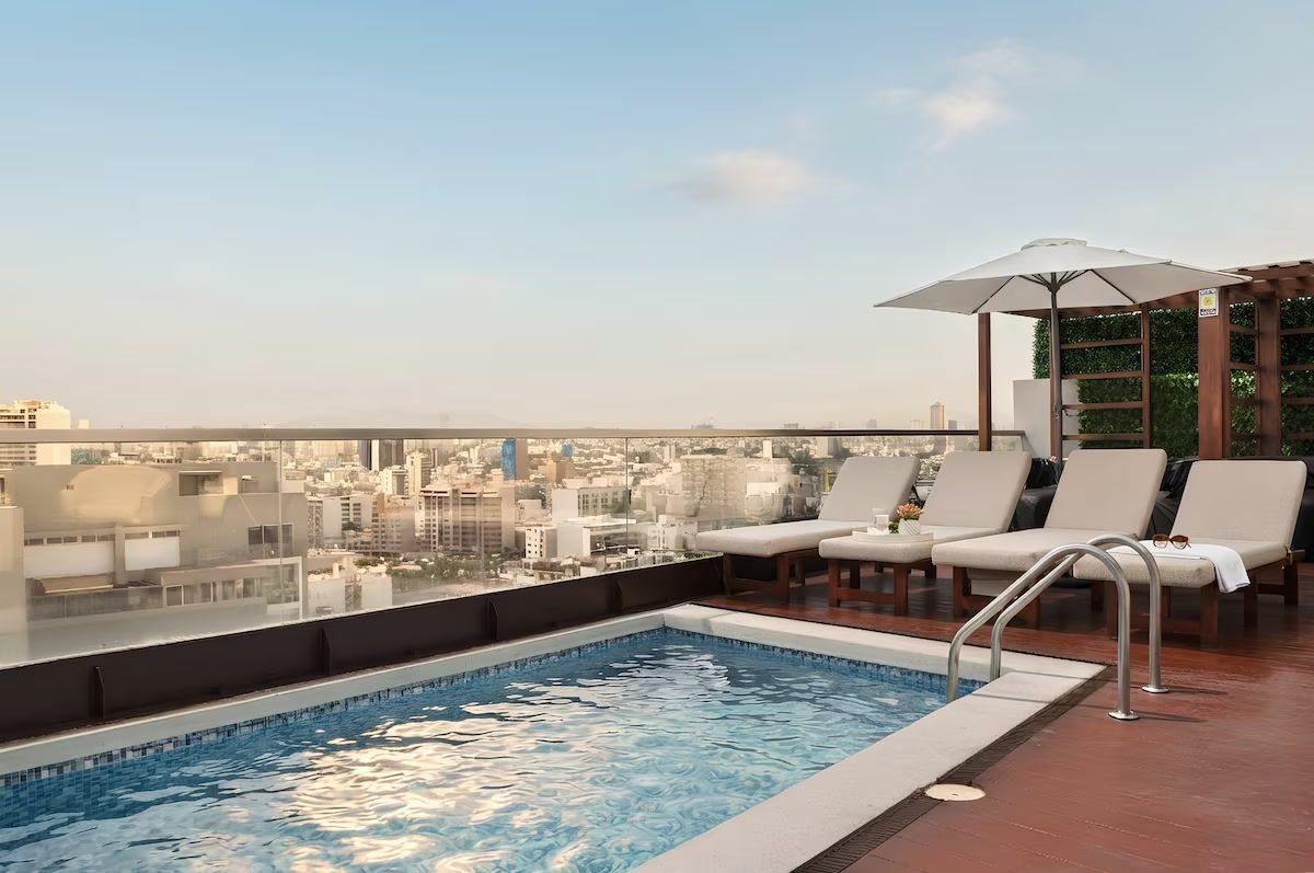 Una piscina en la azotea con sillones de descanso y una sombrilla, con vistas a un paisaje urbano.