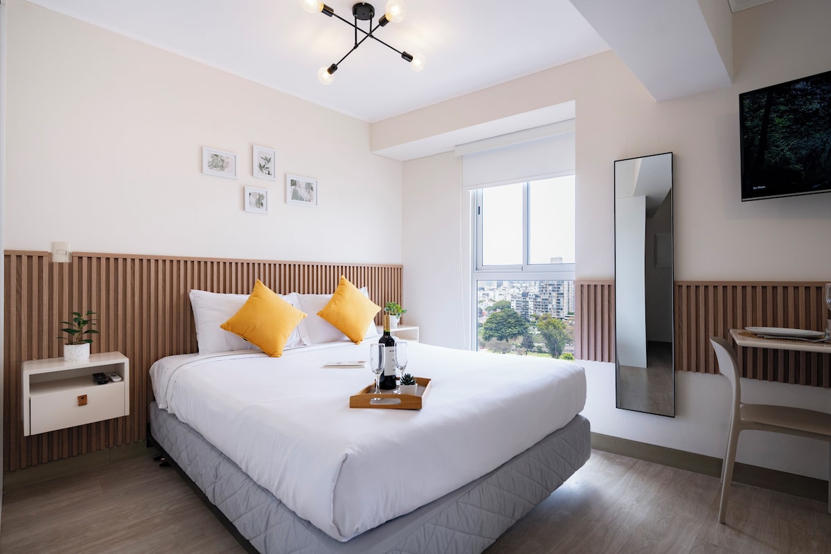 Ein modernes Hotelzimmer mit einem großen Bett, einem Nachttisch, einem Schreibtisch, einem Stuhl, einem Fernseher und einem Fenster mit Blick auf die Stadt.