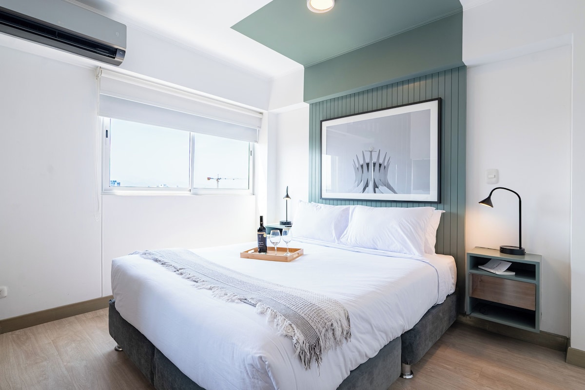 Ein modernes Schlafzimmer mit einem großen Bett, einem Fenster, einem gerahmten Kunstwerk, einem Nachttisch mit Lampe und einer Ablage mit Flasche und Gläsern.