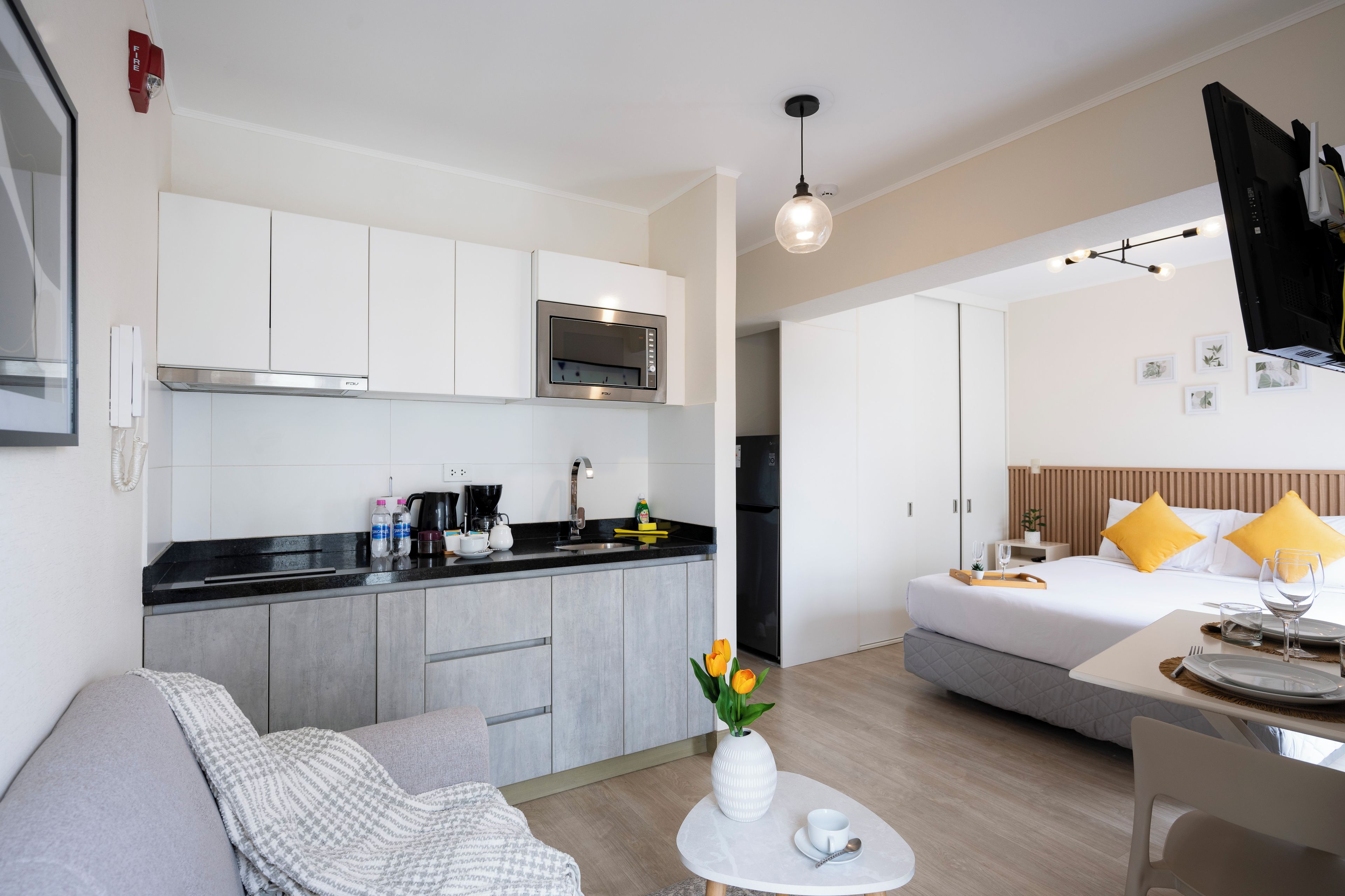 Una habitación de hotel moderna con kitchenette, una cama con almohadas amarillas, zona de comedor y una pequeña mesa redonda con un florero.