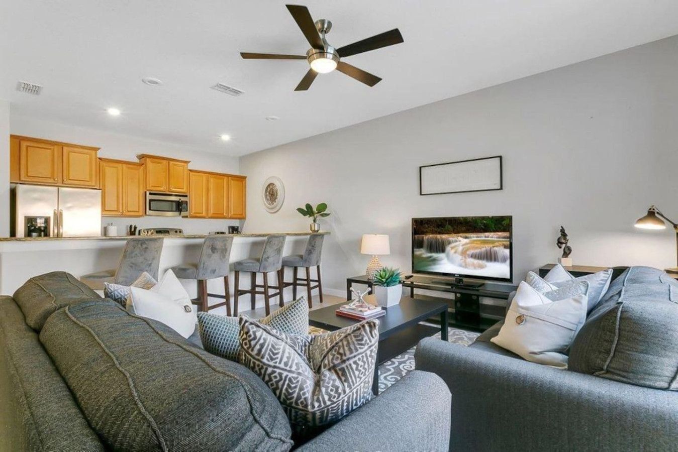 A modern living room with a sofa set, a dining area with bar stools, a kitchen with wooden cabinets, and a ceiling fan.