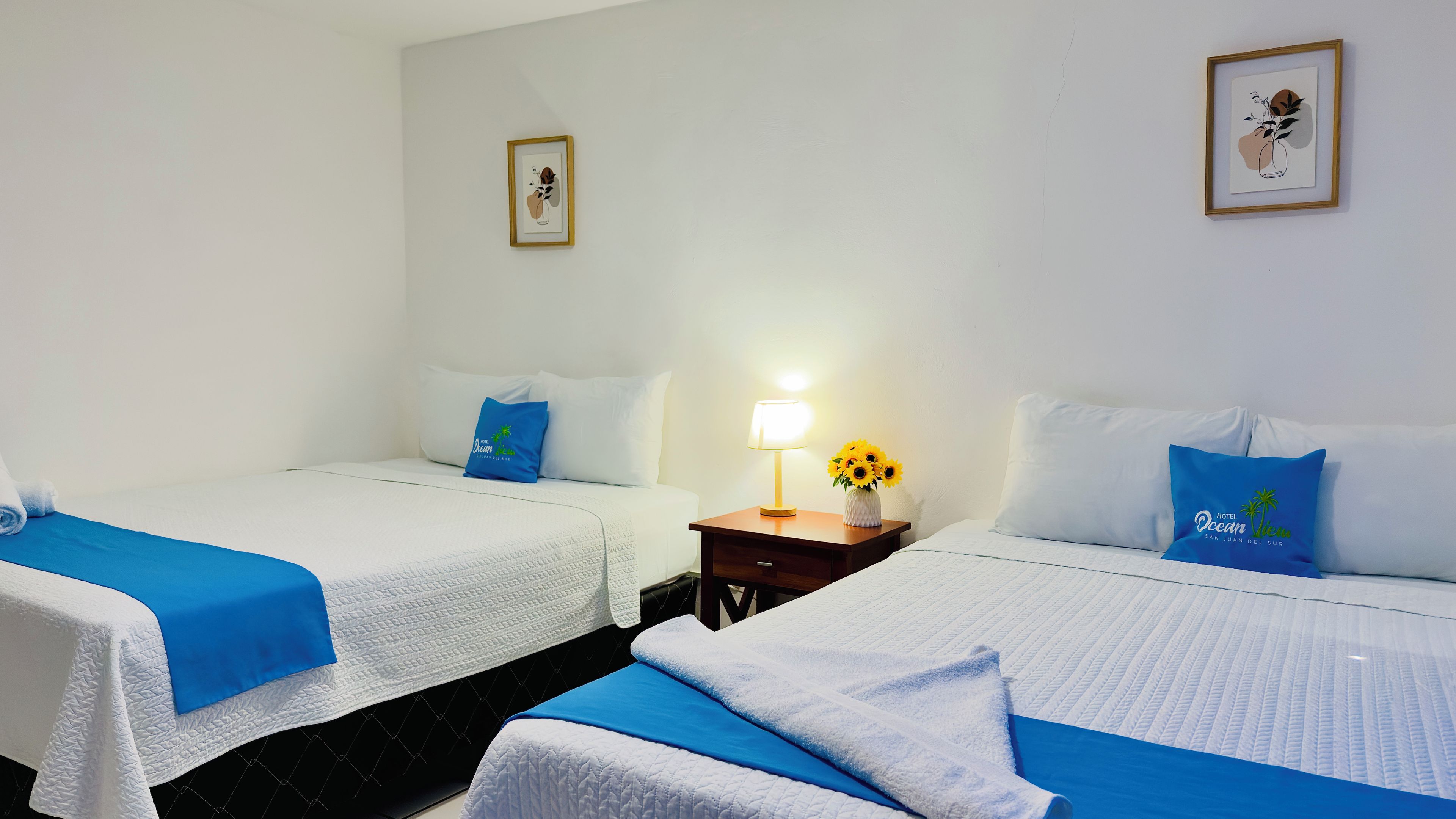Una habitación de hotel con dos camas, una mesita de noche con lámpara y un cuadro en la pared.