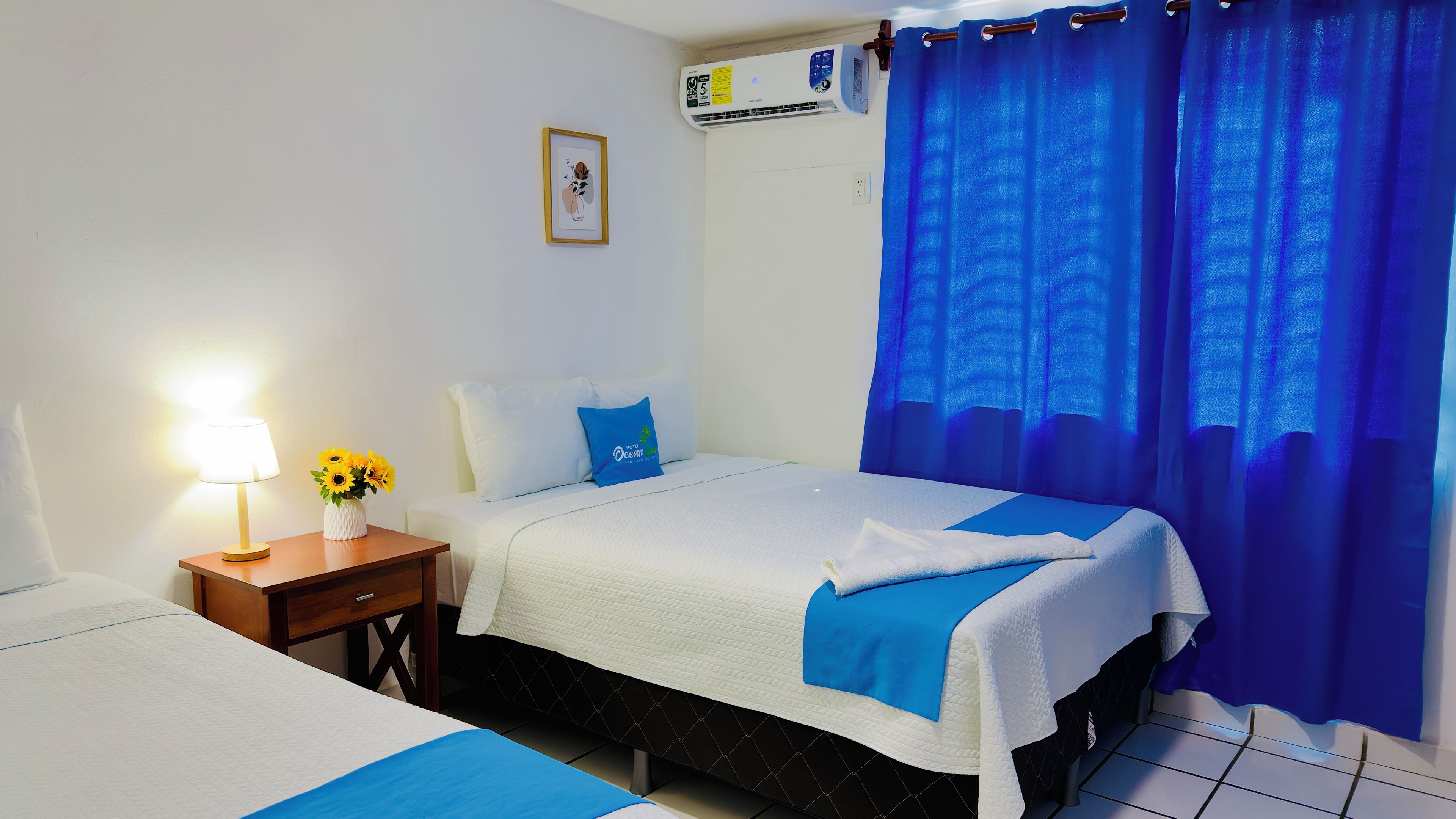 Una habitació d'hotel amb un llit, una taula de nit amb una làmpada i flors, una finestra amb cortines blaves i un aparell d'aire condicionat.