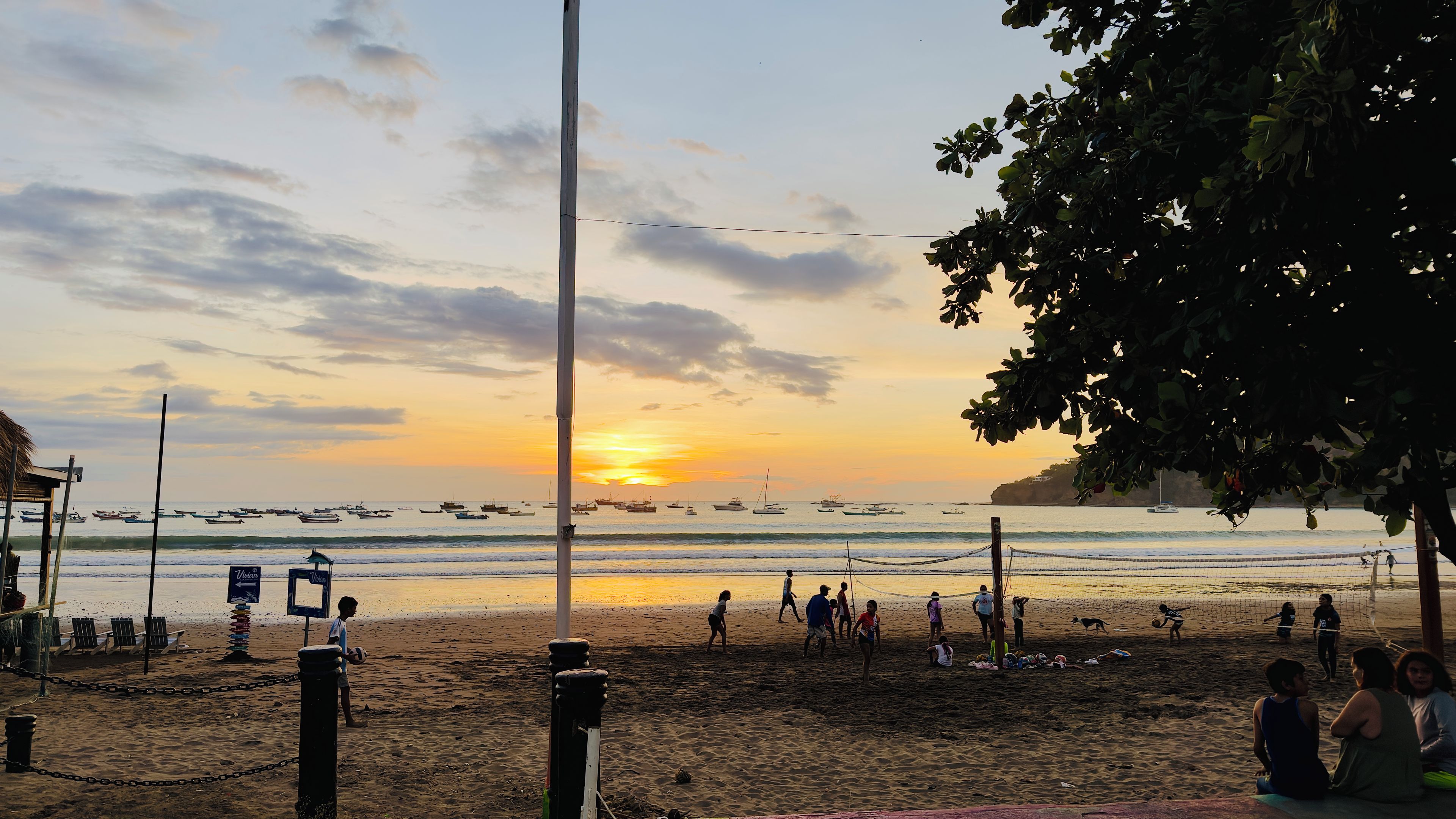 Une plage avec un filet de volley-ball et des gens qui jouent, un coucher de soleil en arrière-plan et des bateaux sur l’eau.
