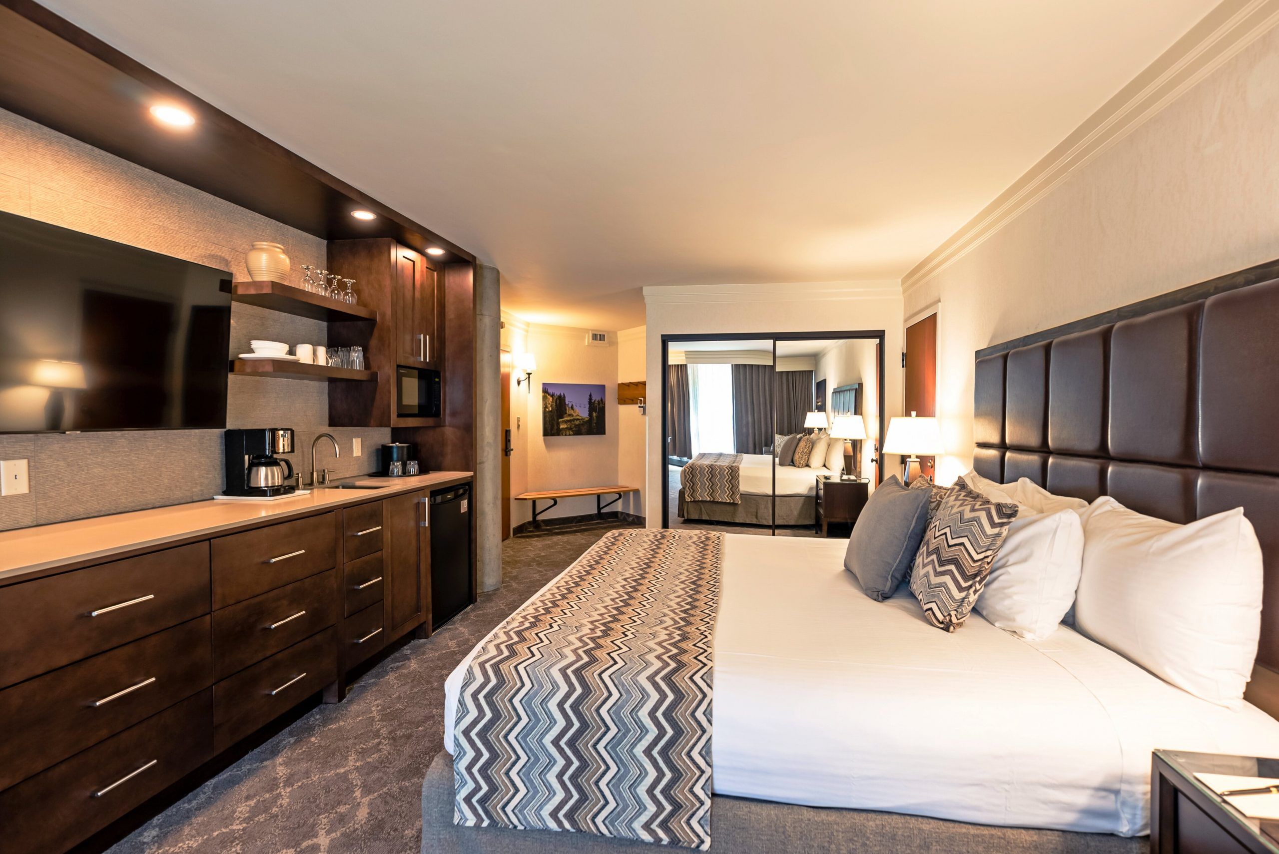 A hotel room with a bed, a bedside table, a lamp, a TV, a desk, and a kitchenette.