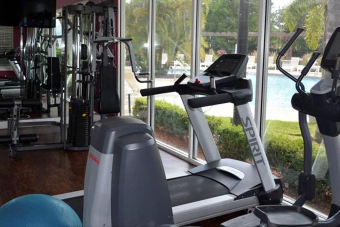 A gym with cardio equipment, including a treadmill and stationary bikes, with a view of a pool and greenery outside.