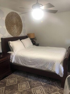 Ein Schlafzimmer mit einem Bett, einem Nachttisch, einer Wanddekoration, einem Deckenventilator und einem gemusterten Teppich.