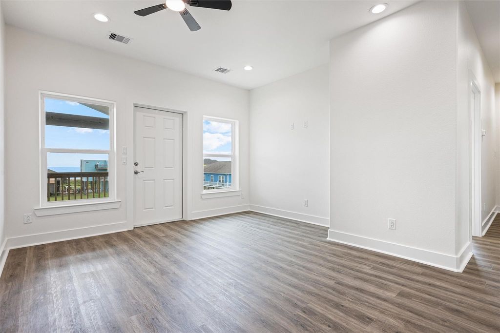 A spacious room with hardwood flooring, two windows with views, a ceiling fan, and recessed lighting.