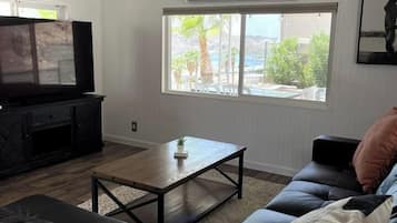 A living room with a black leather sofa, a wooden coffee table, a flat-screen TV on a black entertainment unit, and a large window with a view of greenery and a pool.