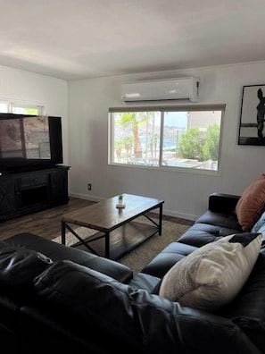 Una sala de estar con un sofá de cuero negro, una mesa de centro de madera, un televisor de pantalla plana sobre un mueble negro y un ventanal con vistas a zonas verdes y una piscina.