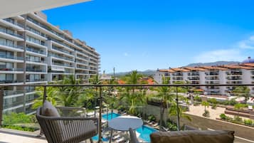 Un balcón con mobiliario de mimbre, una piscina y vistas a una zona residencial con edificios y montañas.
