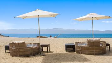 A beach with two wicker lounge chairs under umbrellas, a person relaxing on the sand, and a view of the sea and distant mountains.