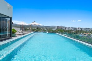 A rooftop infinity pool with clear blue water, surrounded by lounge chairs and umbrellas, overlooking a cityscape with mountains in the distance. - 1BR Condo in La Cruz de Huanacaxtle With Pool, Gym, Wifi, and AC (La Cruz de Huanacaxtle)