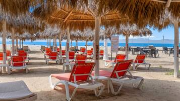 Un espace de détente aménagé sur la plage, avec des rangées de chaises longues blanches et rouges disposées sous des parasols en chaume, offrant une vue sur la mer.