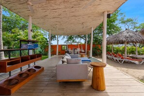 A covered outdoor seating area with a sofa, wooden tables, and a thatched umbrella. - Oceanfront 1BR Condo With Pool, Gym, AC, and Balcony (La Cruz de Huanacaxtle)