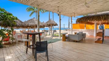 Un espace extérieur aménagé, abrité et doté de tables, de chaises, d’un canapé, et offrant une vue sur la plage et l’océan.