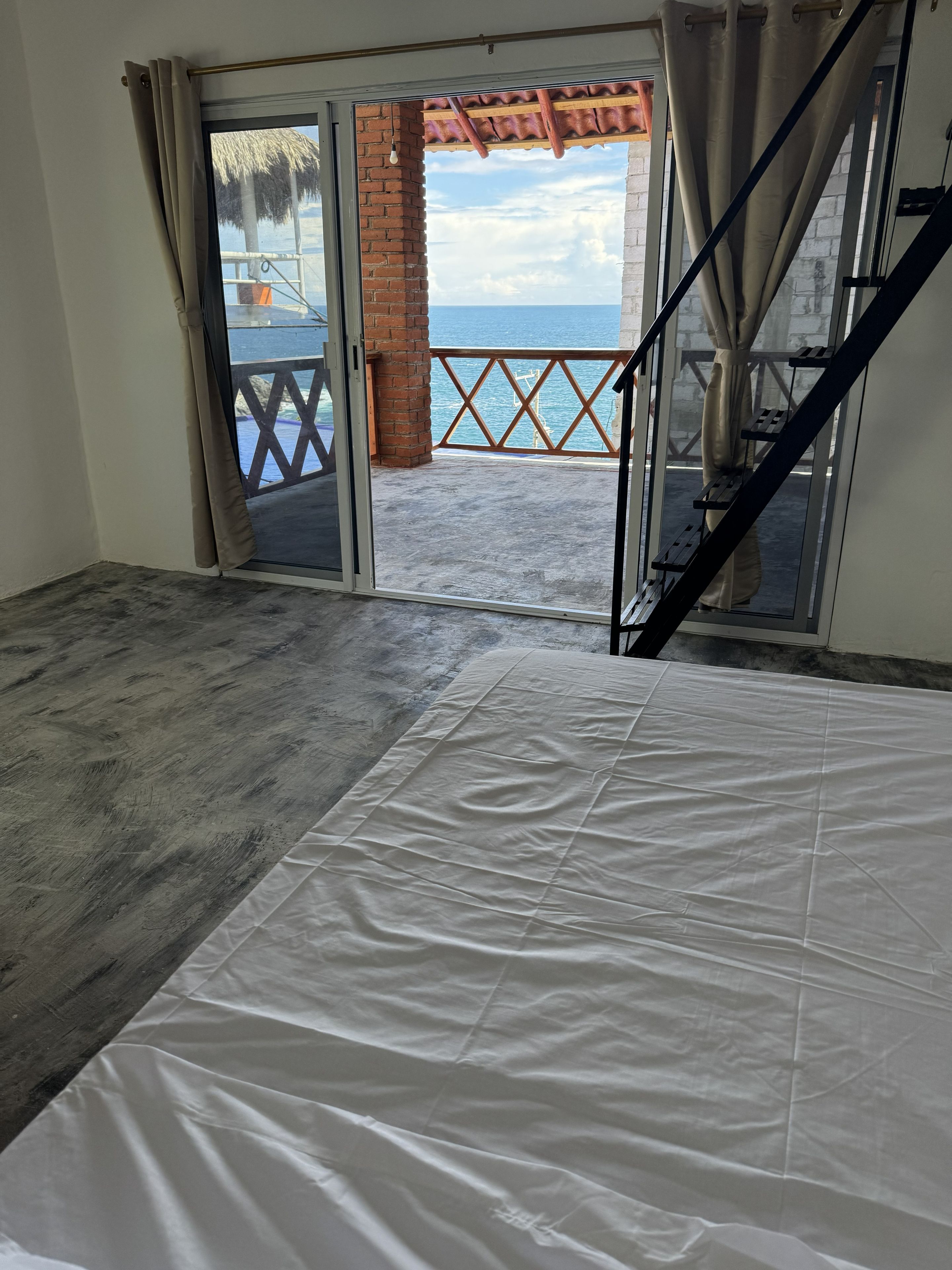 Habitación con una cortina blanca grande, vista al mar a través de un vidrio y un pilar de ladrillo, además de una barandilla de madera.