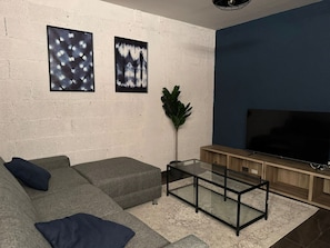 Un soggiorno con un divano componibile, un tavolino in vetro, una televisione a schermo piatto e due opere d'arte incorniciate appese al muro.