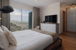 A bedroom with a large bed, a TV, and a view of the city. - Casa do Pedregal – Sea Views in Câmara de Lobos (Funchal)