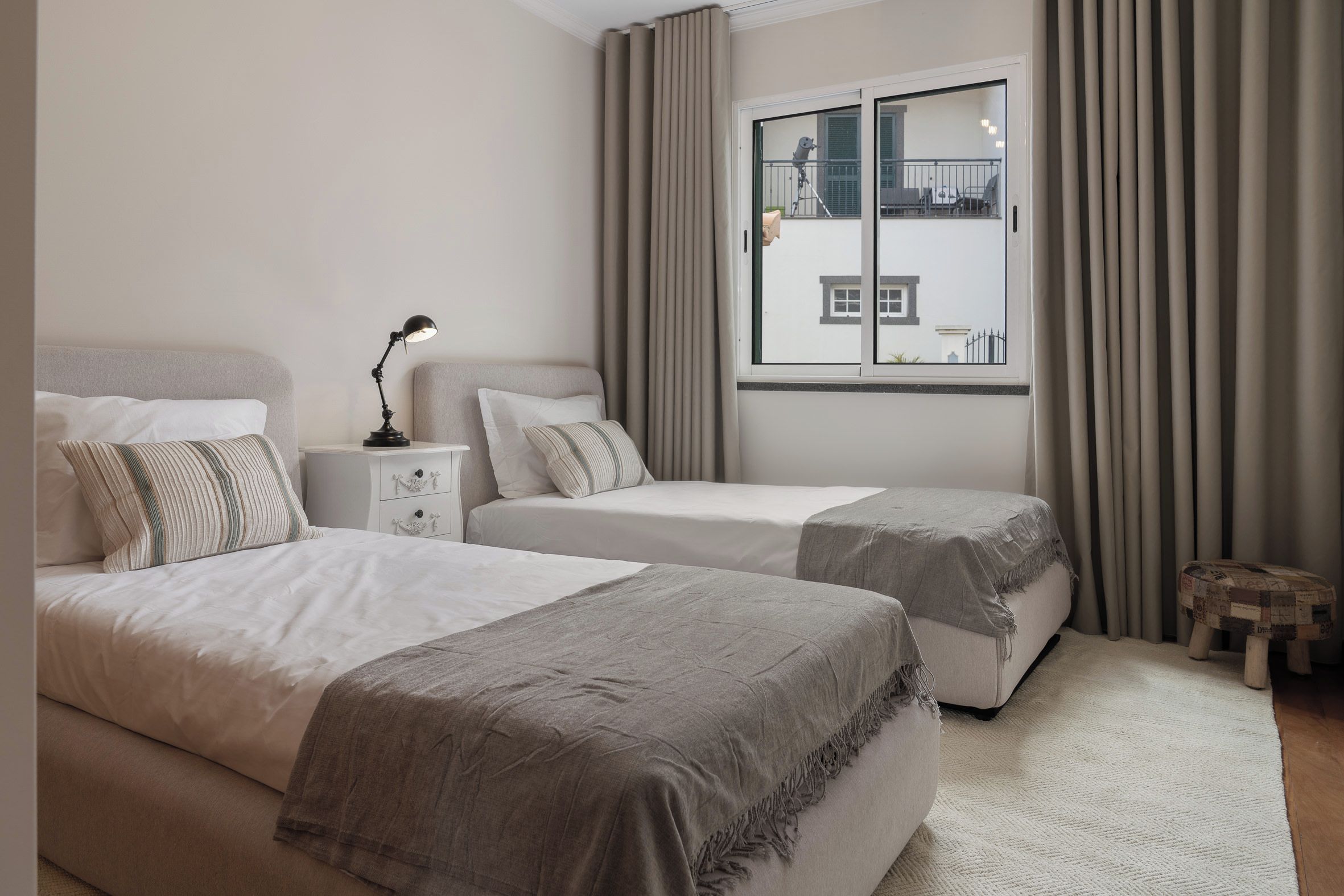 Une chambre d’hôtel avec deux lits, une table de chevet, une lampe, une fenêtre avec des rideaux et un petit tabouret.