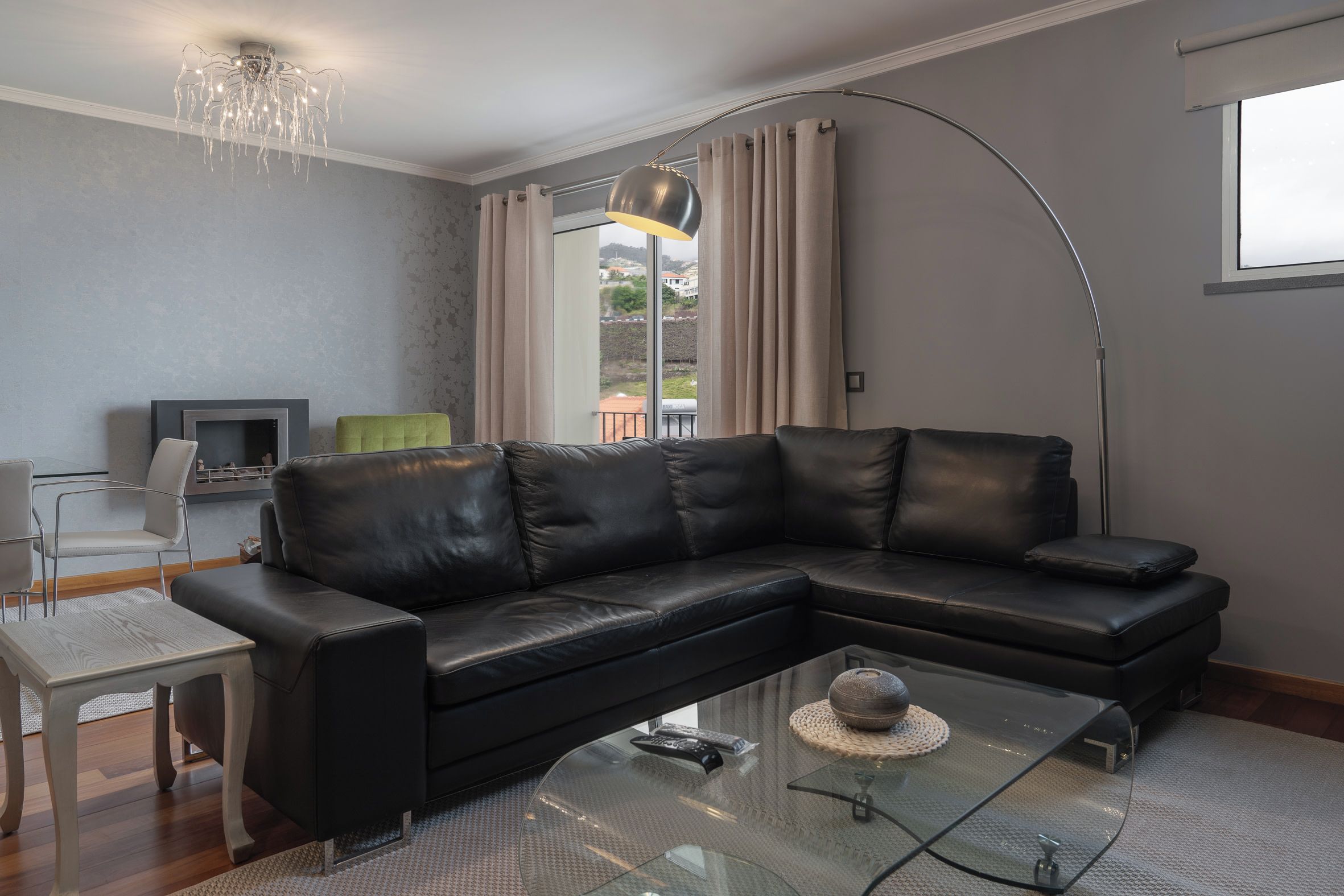 Un salon moderne avec un canapé d’angle en cuir noir, une table basse en verre, une table à manger avec des chaises, un téléviseur à écran plat et une grande fenêtre avec des rideaux.