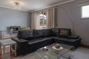 A modern living room with a black leather sectional sofa, a glass coffee table, a dining table with chairs, a flat-screen TV, and a large window with curtains. - Casa do Pedregal – Sea Views in Câmara de Lobos (Funchal)