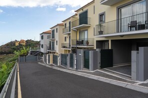 A residential building with balconies, a gated entrance, and a view of a hill with a building in the distance.