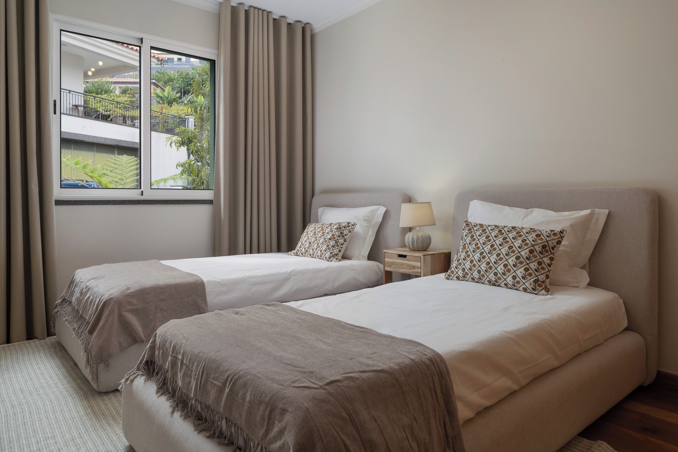 A hotel room with two beds, a window with curtains, a bedside table with a lamp, and a view of greenery outside.