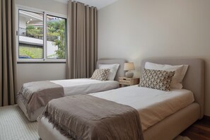 A hotel room with two beds, a window with curtains, a bedside table with a lamp, and a view of greenery outside.