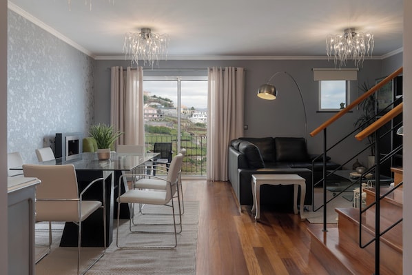 A modern living room with a dining area, a black leather sofa, a staircase, and a view of the city.