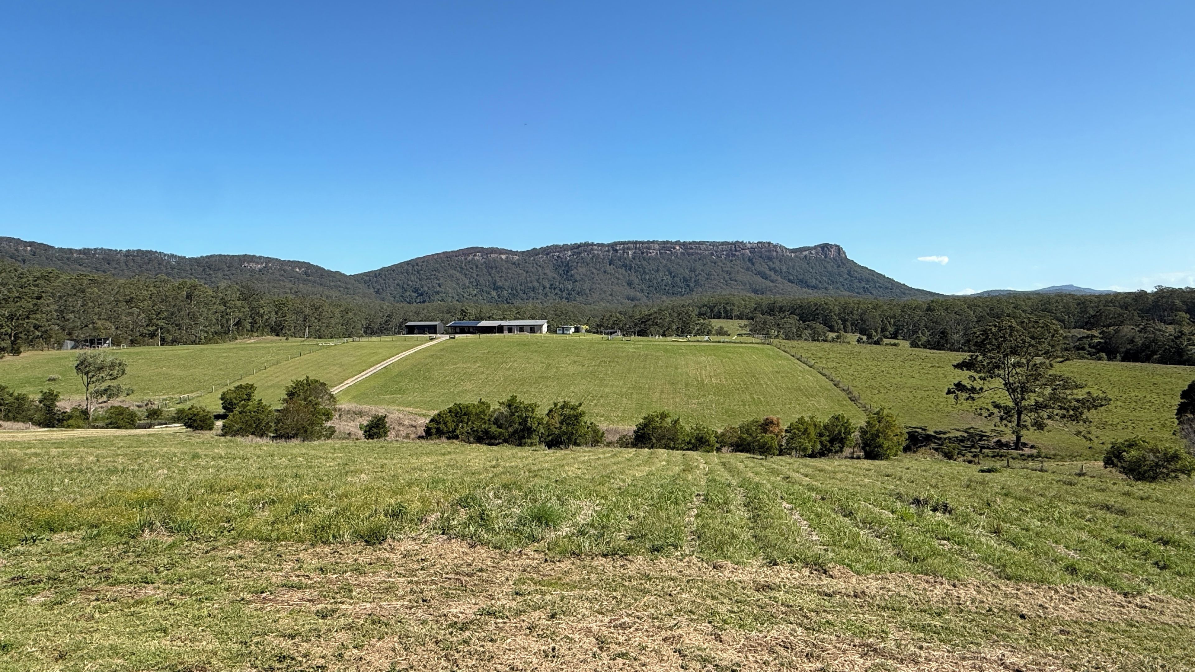 A rural landscape with a large open field, a building with a flat roof, and a mountain in the background.