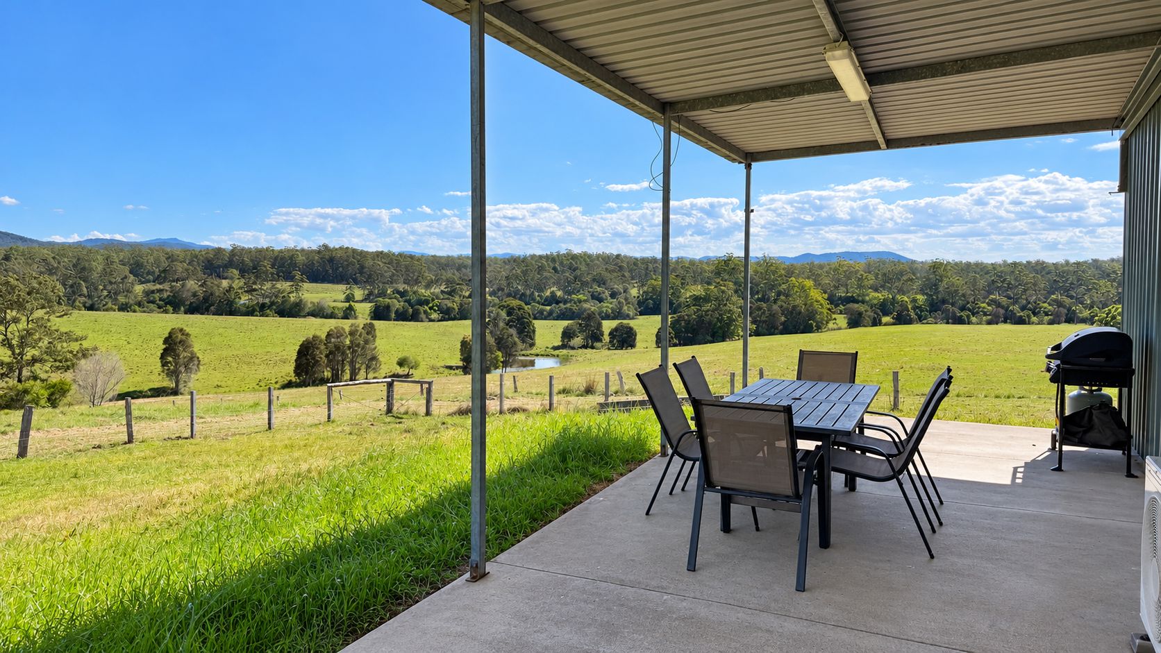 An outdoor patio with a table and chairs, a barbecue grill, and a view of a green field and mountains.