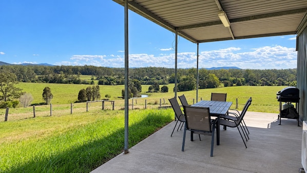 Un patio exterior con mesa y sillas, una parrilla de barbacoa y vistas a un campo verde y montañas.