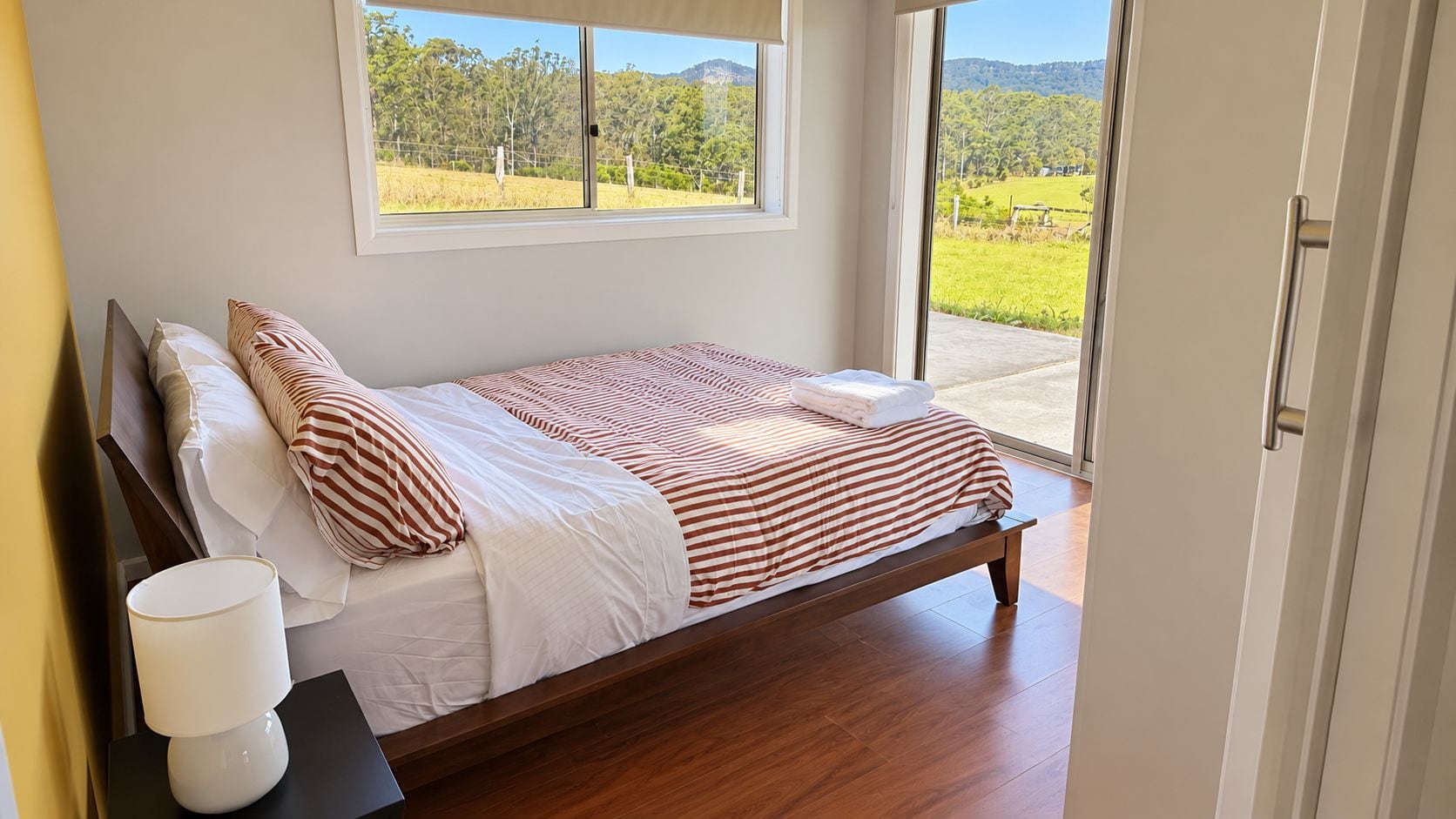 A single bed with a wooden frame, white bedding, and striped pillows, positioned next to a window with a view of a green landscape.