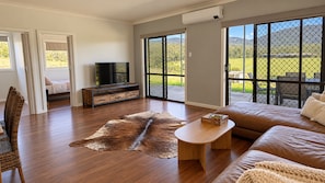 A spacious living room with a large sofa, a coffee table, and a television. There is a sliding glass door leading to an outdoor area with a view of the countryside.