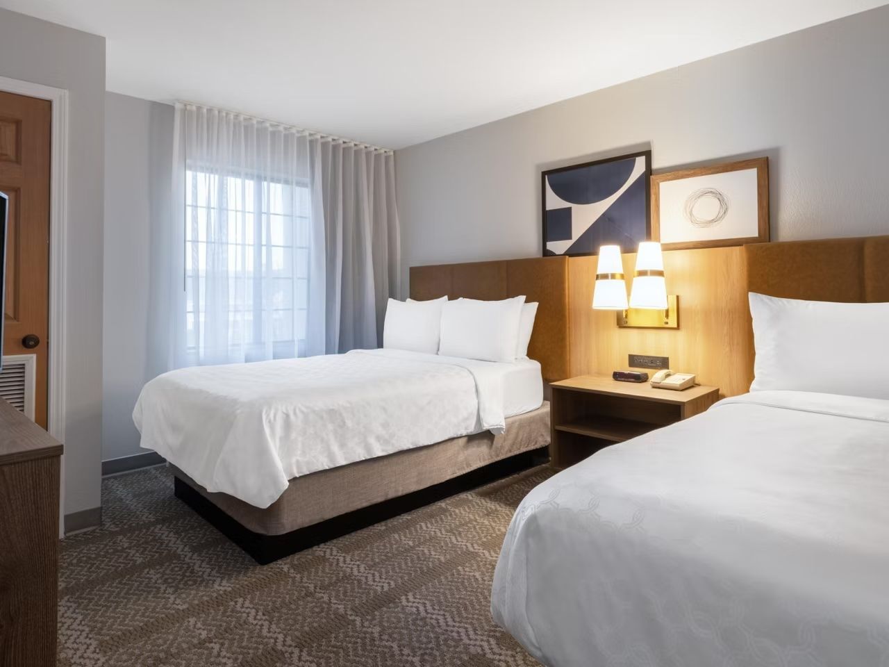 A hotel room with two beds, a nightstand with a lamp, a window with curtains, and a wall-mounted artwork.