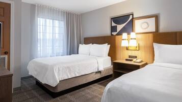 A hotel room with two beds, a nightstand with a lamp, a window with curtains, and a wall-mounted artwork.