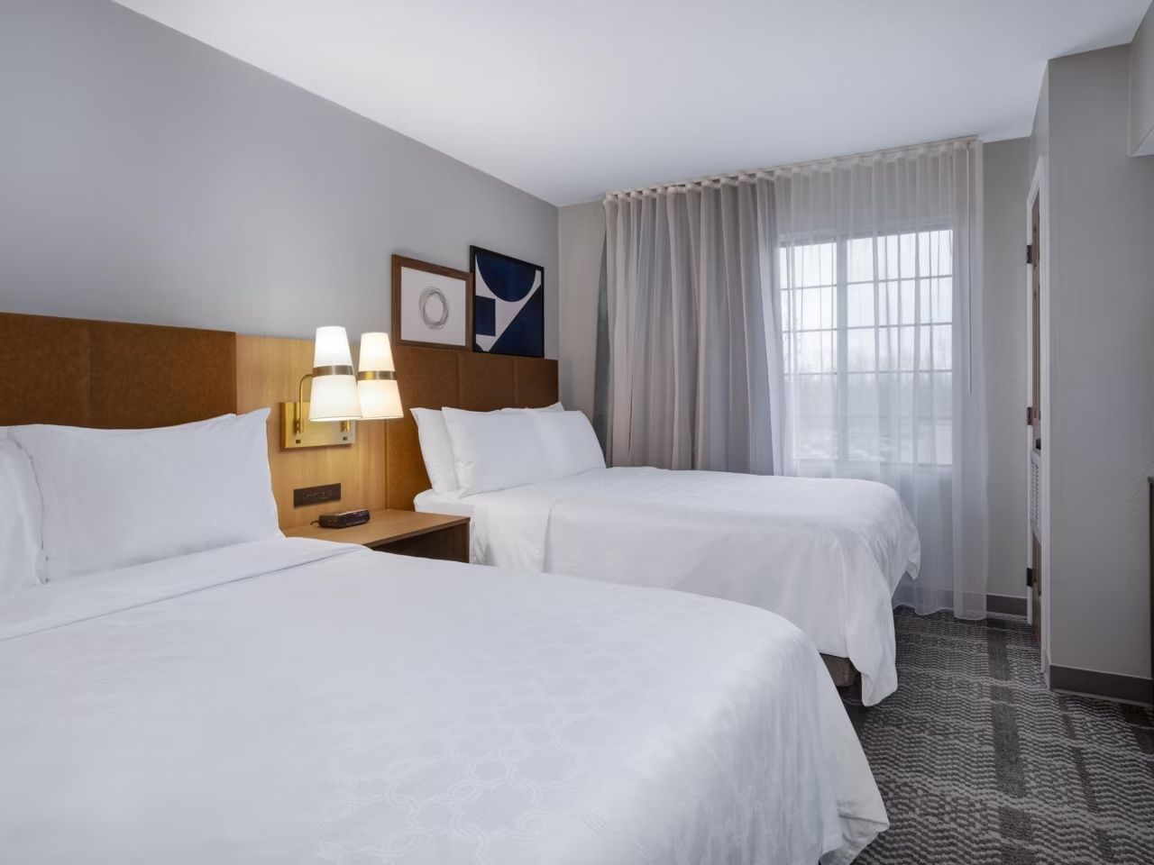 A hotel room with two beds, a nightstand with a lamp, a window with sheer curtains, and a wall-mounted picture.