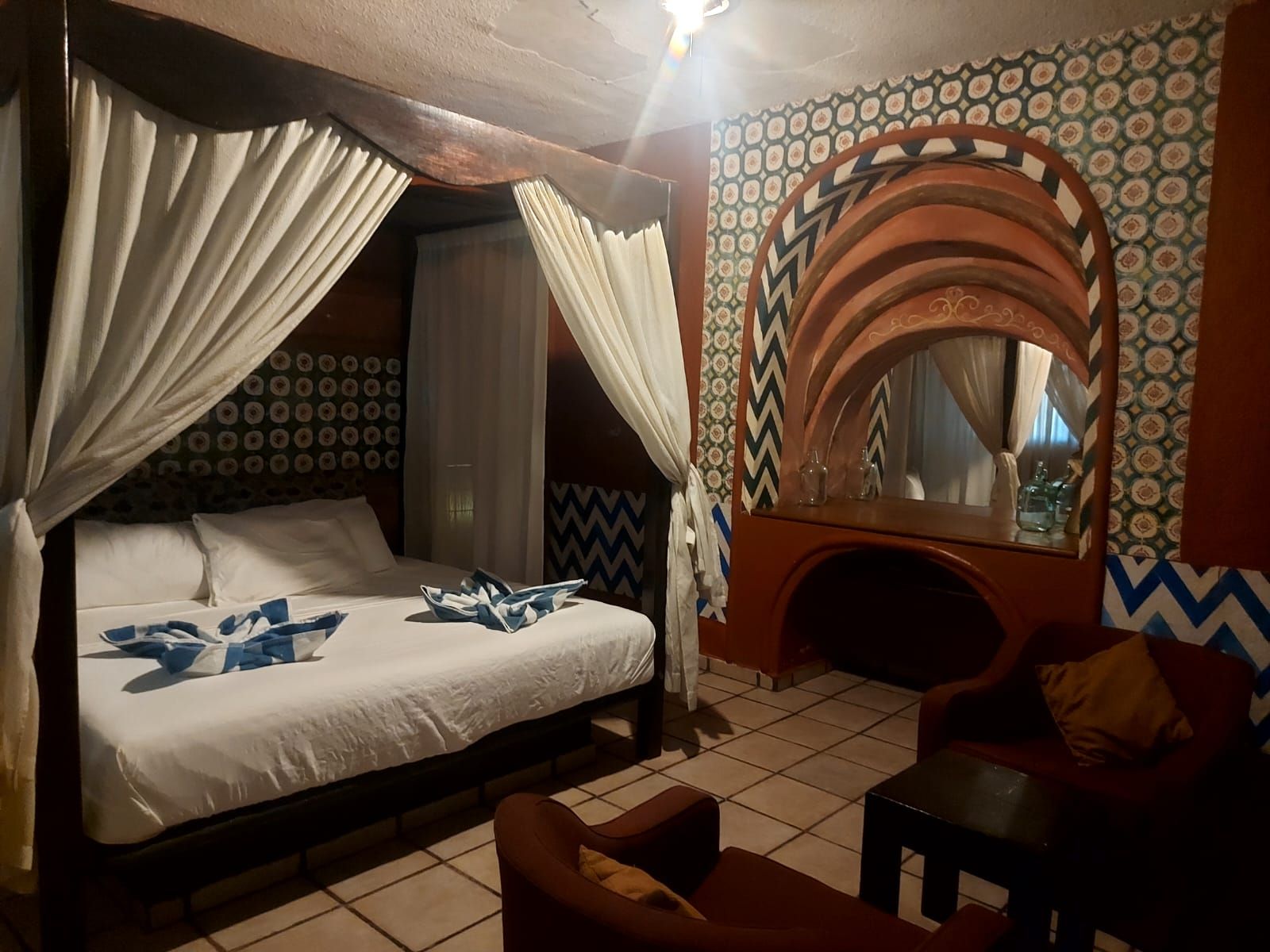 Quarto de hotel com cama, área de estar com poltrona e mesa pequena, espelho decorativo e papel de parede estampado.