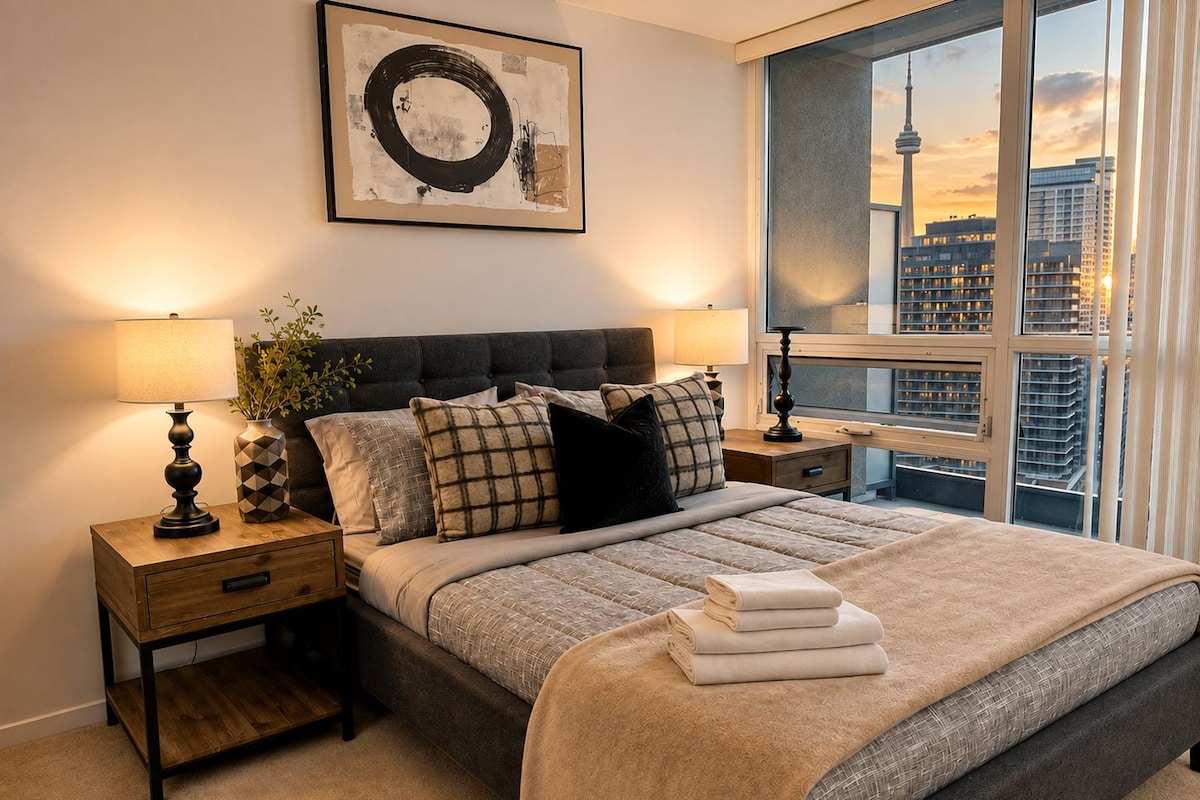 Un dormitorio moderno con una cama grande, dos mesitas de noche, vistas a la ciudad y una obra de arte enmarcada en la pared.