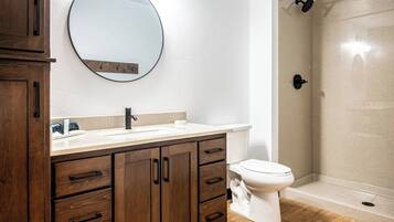 Un baño con tocador de madera, espejo redondo, inodoro y cabina de ducha con mampara de vidrio.
