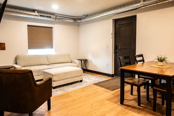 Une pièce comprenant un canapé, une table à manger avec des chaises, une petite table ronde, une porte en bois foncé et une fenêtre avec des stores.